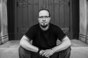 A black and white photo of a man sitting in front of a door taken as part of the Peter Jennings Disciple's album art for their single "Not Enough Bridge.