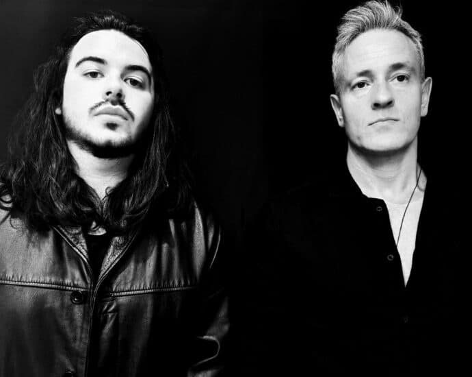 In a striking black and white photo, one man with long hair and a leather jacket stands beside another with short hair in a collared shirt. Their distinct styles create an elementor of contrast against the dark background.